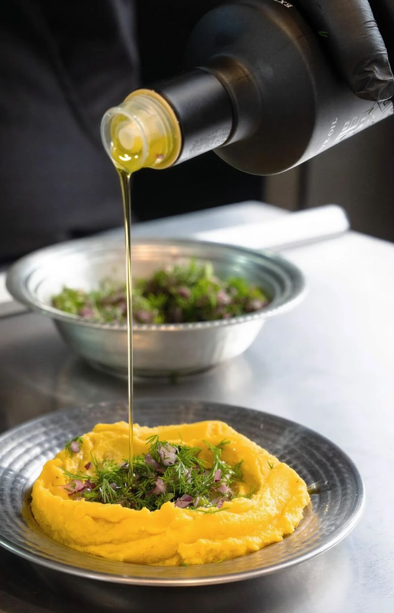Oro di Milas olive oil poured from the bottle by a gloved hand over a yellow bean purée topped with dill and red onion, a bowl of herbs behind